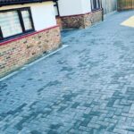 Grey block paving driveway in Basildon featuring a herringbone pattern and red brick wall.